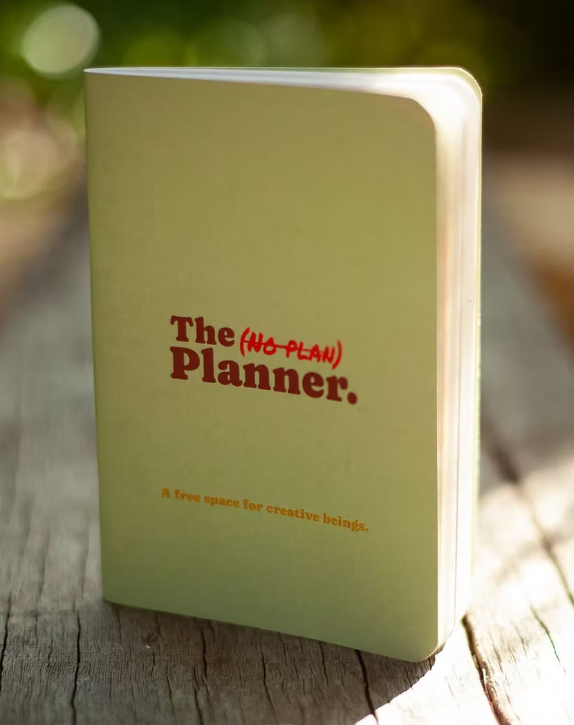 Olive green mini notebook titled 'The (No-Plan) Planner' on a wooden surface.