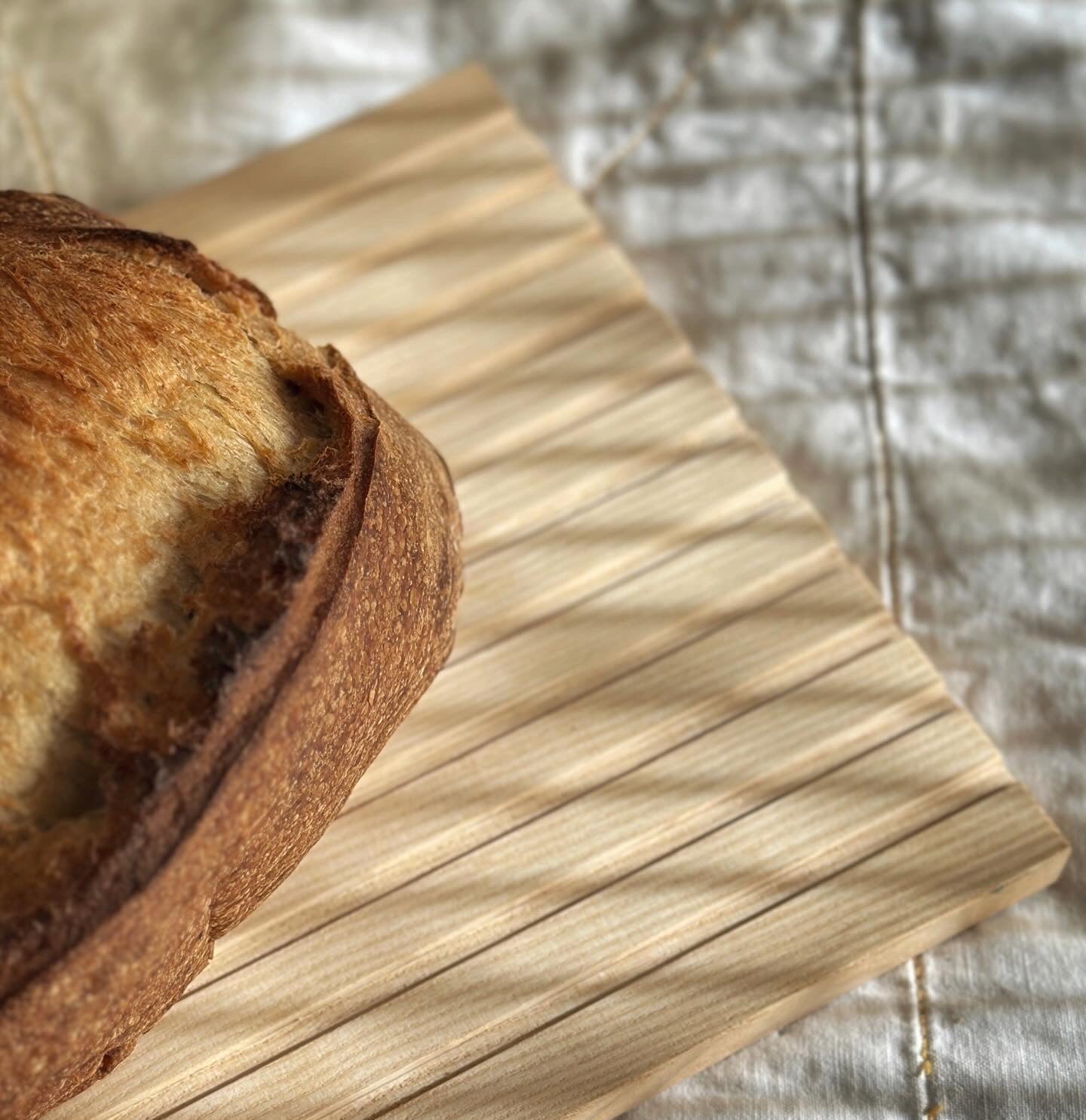 A close-up of a loaf of bread on a rectangular timber cutting board with a ridged texture, placed on a textured beige fabric background. 